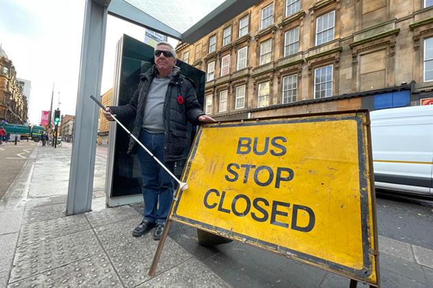 Sandy Taylor, NFBUK Chair Scotland, holding his white cane standing in a bus shelter on Sauchiehall Street 